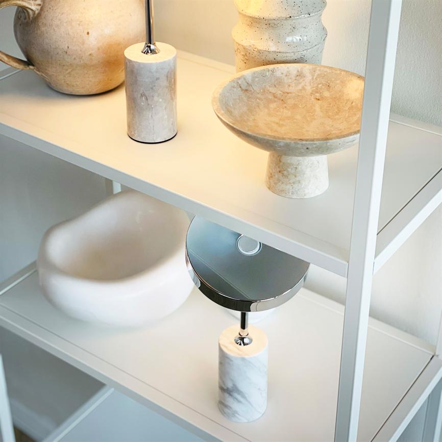 Marmorbaseret bordlampe står og reflekterer blødt lys ved siden af en lav sten- eller keramikskål og en rund kande, arrangeret på hvide metalhylder i et lyst, minimalistisk rum.