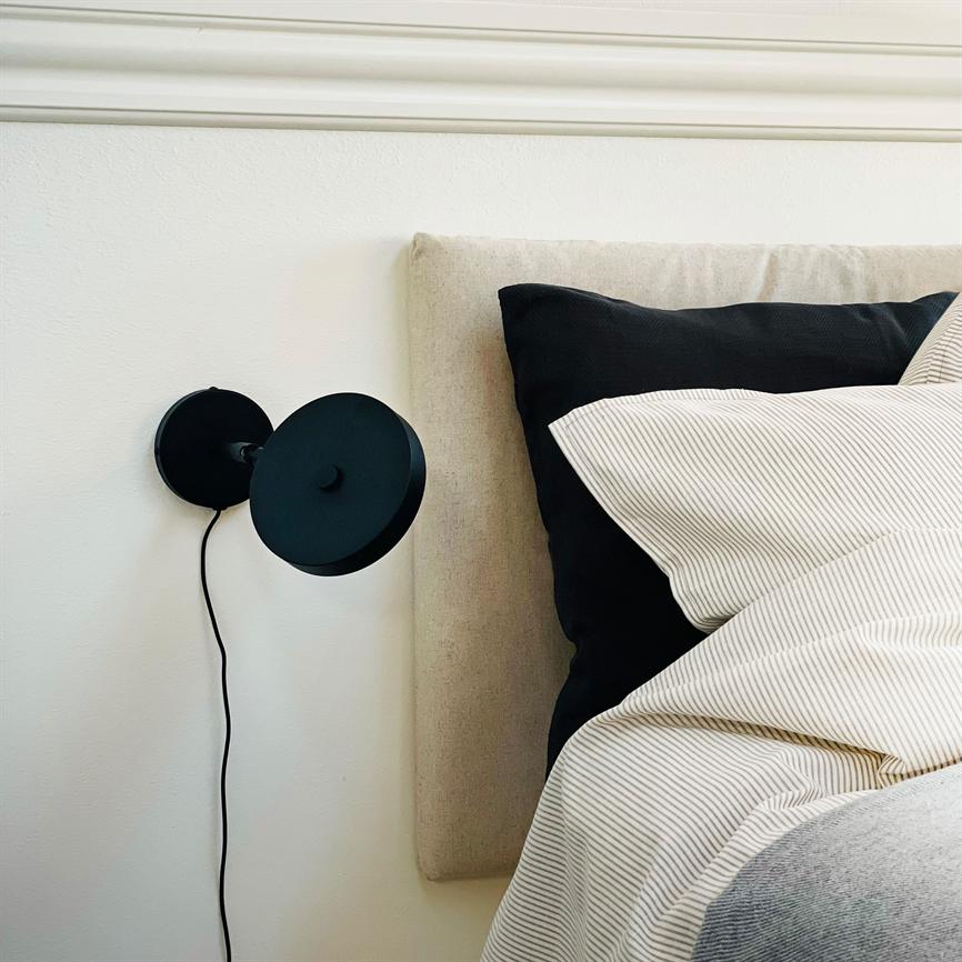 En sort væglampe er monteret på væggen og peger mod en lys, polstret sengegavl; ved siden af ligger en sort pude og en cremehvid, tyndstribet dyne.