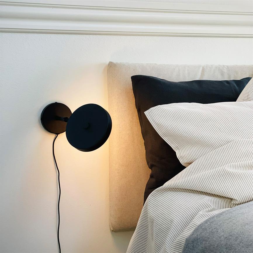 En sort væglampe lyser blødt ved en beige sengegavl med sort ledning; en mørk pude og hvidt, fintstribet sengetøj ligger på en seng i et lyst, minimalistisk soveværelse.