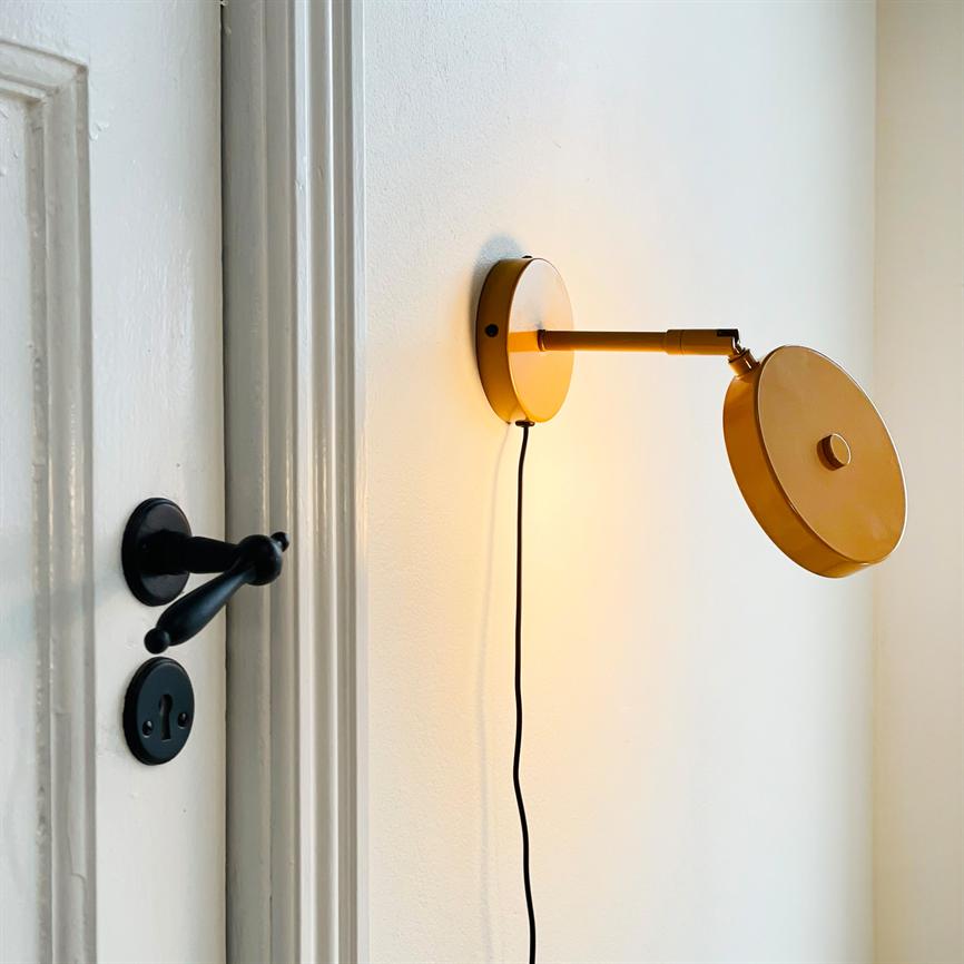 Gul væglampe monteret på hvid væg, arm udstrakt med skiveformet skærm vippet; sort ledning hænger ned; ved siden en hvid dør med sort dørhåndtag og nøgleskilt.