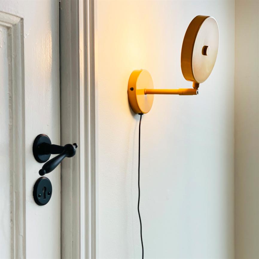 En gul væglampe lyser, monteret med en rund skærm og en ledning hængende ned; den sidder på en hvid væg ved siden af en hvid dør med sort dørhåndtag.