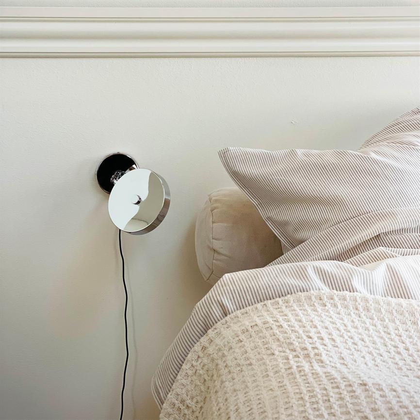 En rund krom væglampe sidder monteret på en hvid væg; dens sorte ledning hænger ned. Ved siden af står en seng med beige rullepude, stribet pudebetræk og lyst, vævet tæppe.