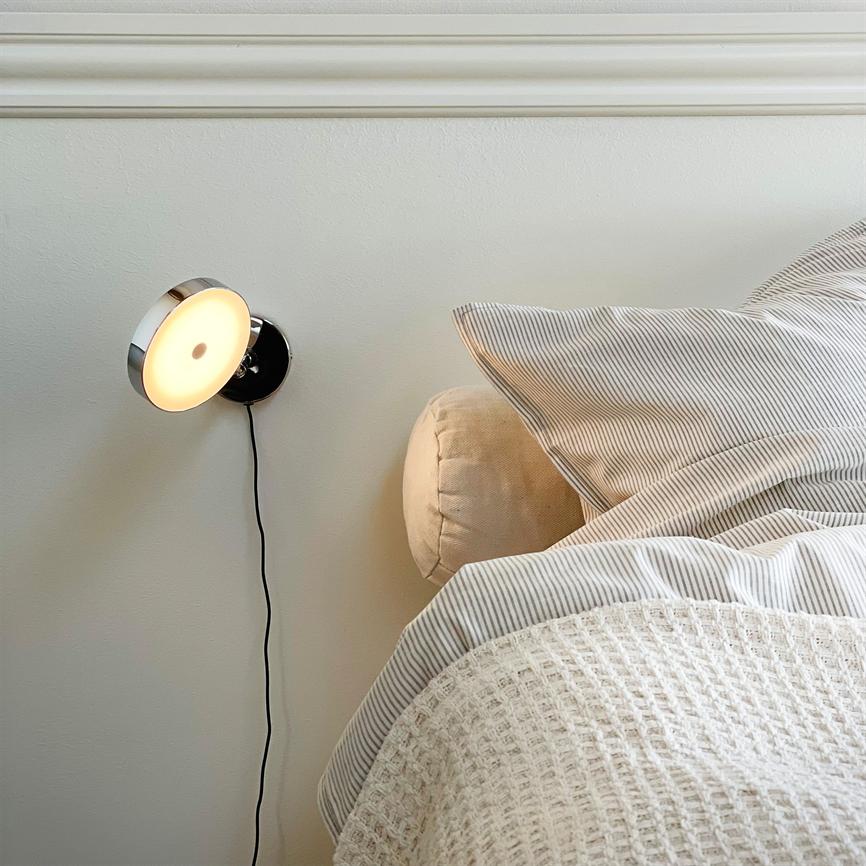 En rund væglampe lyser blødt, dens sorte ledning hænger ned; ved siden af en neutral seng med stribet pude og struktureret tæppe i et lyst soveværelse.