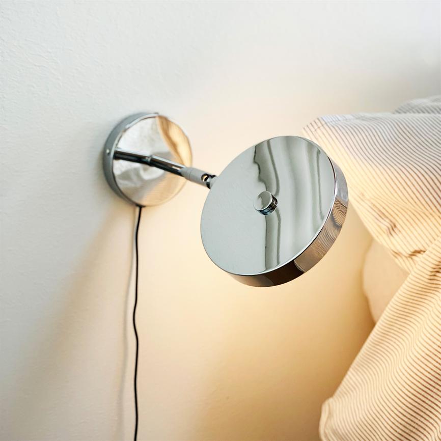 Krom væglampe hænger på en hvid væg, lyser varmt nedad og kaster refleksioner på sin skinnende overflade; den sidder ved en seng med lyse, fintstribede puder.