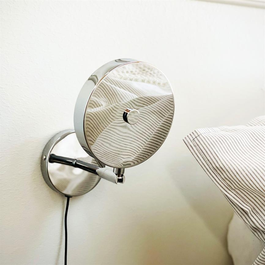 Rund krom vægspejl reflekterer stribet sengetøj; monteret på en arm ved en hvid væg, ved siden af en pude med tynde grå striber.