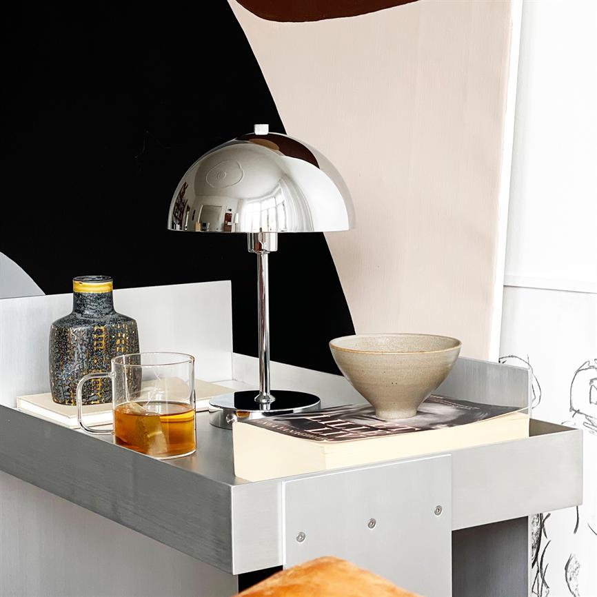 En krom kuppel-lampe står på et sølvfarvet natbord, reflekterer rummet; omkring den et glas med te, en mønstret vase, en skål og en bog foran en abstrakt sort-rosa væg.