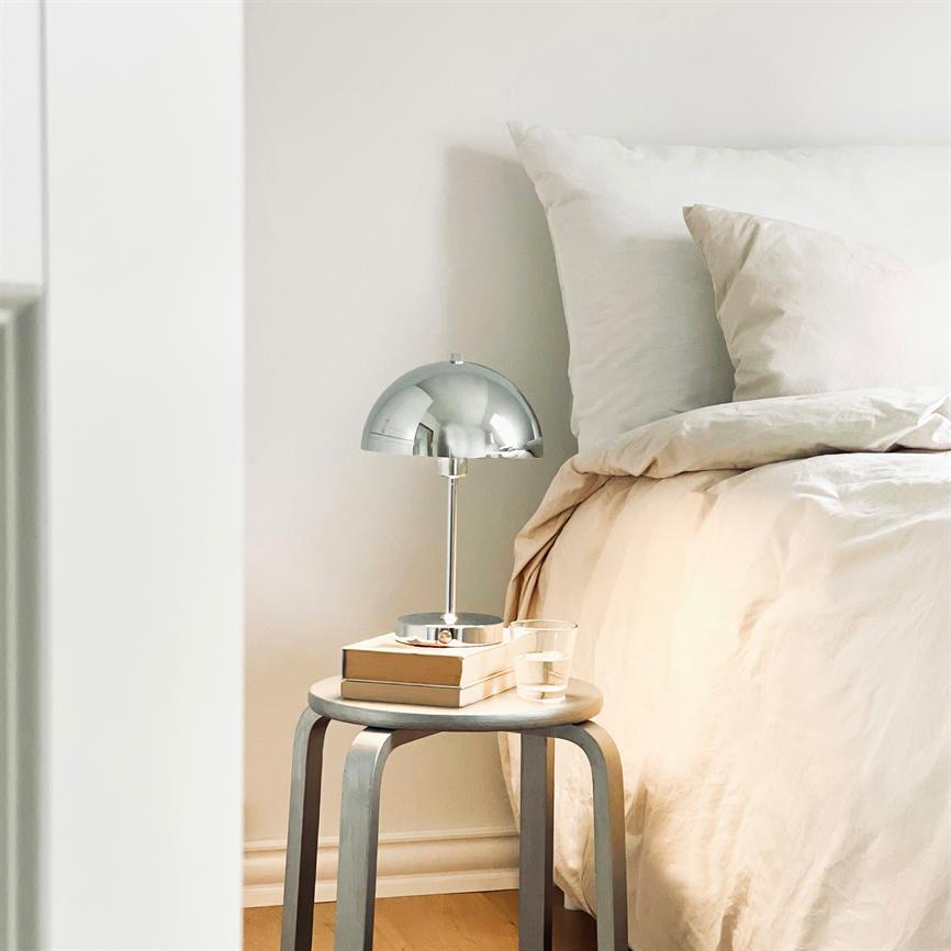 Krom bordlampe står på en rund skammel og lyser svagt over en stak bøger og et glas vand, ved en lysebeige seng med puder i et roligt, minimalistisk soveværelse.