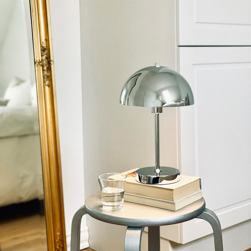 Krom bordlampe står på to bøger på en rund skammel; et glas vand ved siden; i et roligt soveværelseshjørne med et forgyldt spejl og et hvidt skab.