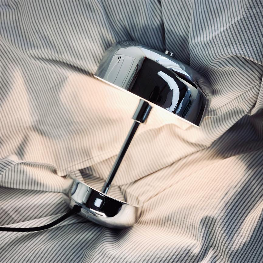 En krom bordlampe ligger på siden; skærmen reflekterer omgivelserne og udsender et blødt lys, placeret på et krøllet, fintstribet lagen.