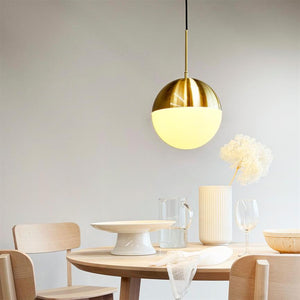 En rund, to-tonet pendellampe hænger og kaster varmt lys over et rundt træspisebord; bordet har et kagefad, glas, høj keramikvase med tørret blomst og en klar vandkande.