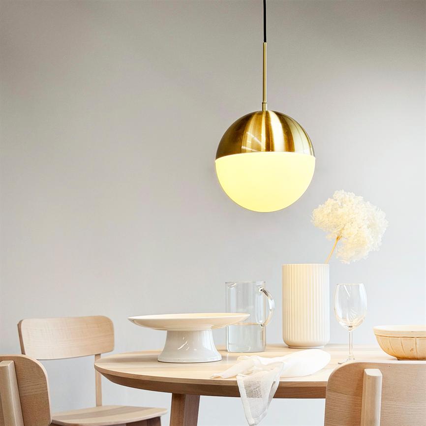 En rund, to-tonet pendellampe hænger og kaster varmt lys over et rundt træspisebord; bordet har et kagefad, glas, høj keramikvase med tørret blomst og en klar vandkande.