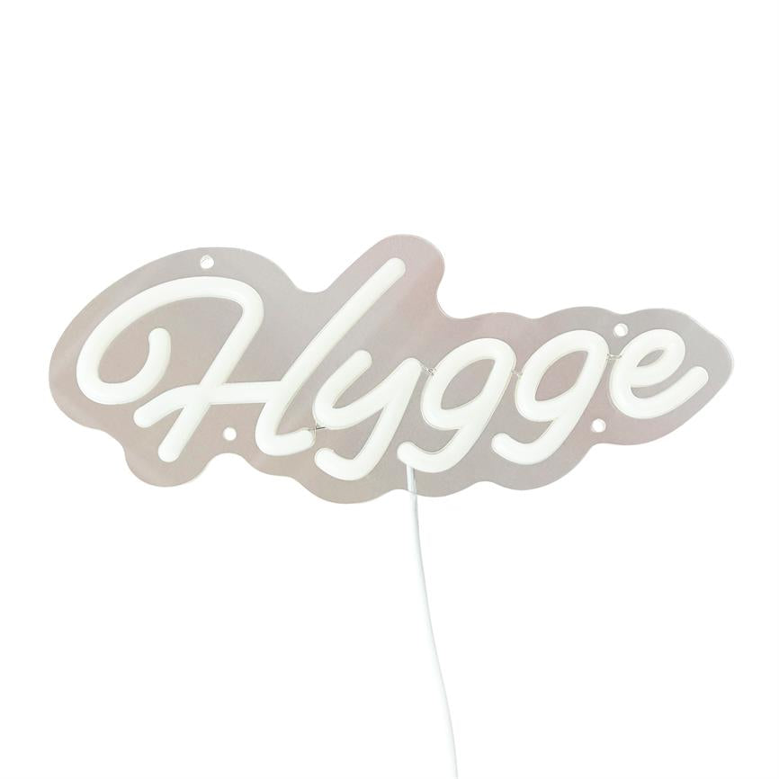 Neonskiltet med teksten "Hygge" lyser blødt, hænger på en hvid baggrund og har en synlig hvid ledning nedad.