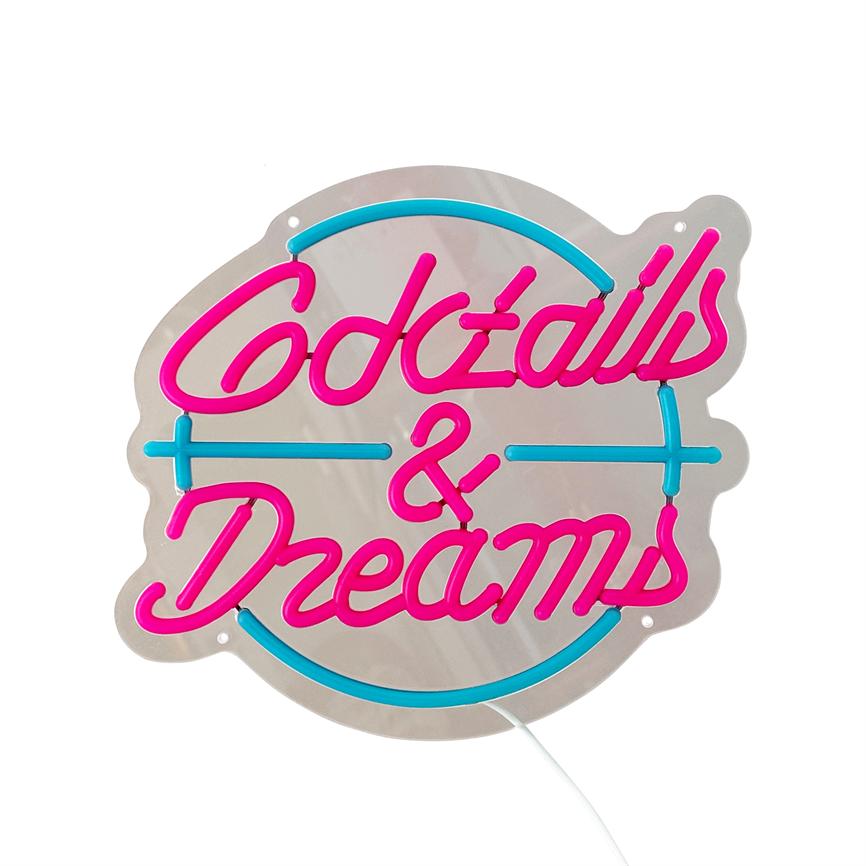 Neonskilt med teksten "Cocktails & Dreams" lyser i pink og blå; bogstaver monteret på en rund, blank metalplade hænger foran en ensartet hvid baggrund med synlig hvid ledning.