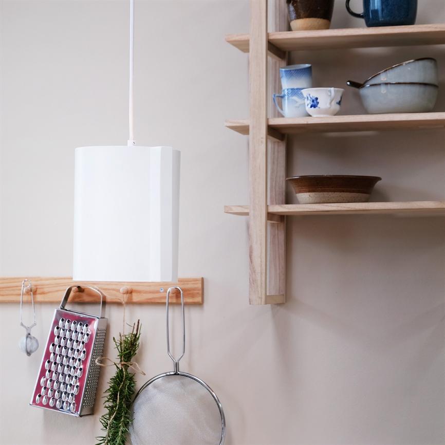 Hvid pendellampe hænger over en træknage; rivjern, en si og et bundt rosmarin hænger fra knagen; til højre åbne træhylder med kopper og skåle i et lyst køkken.