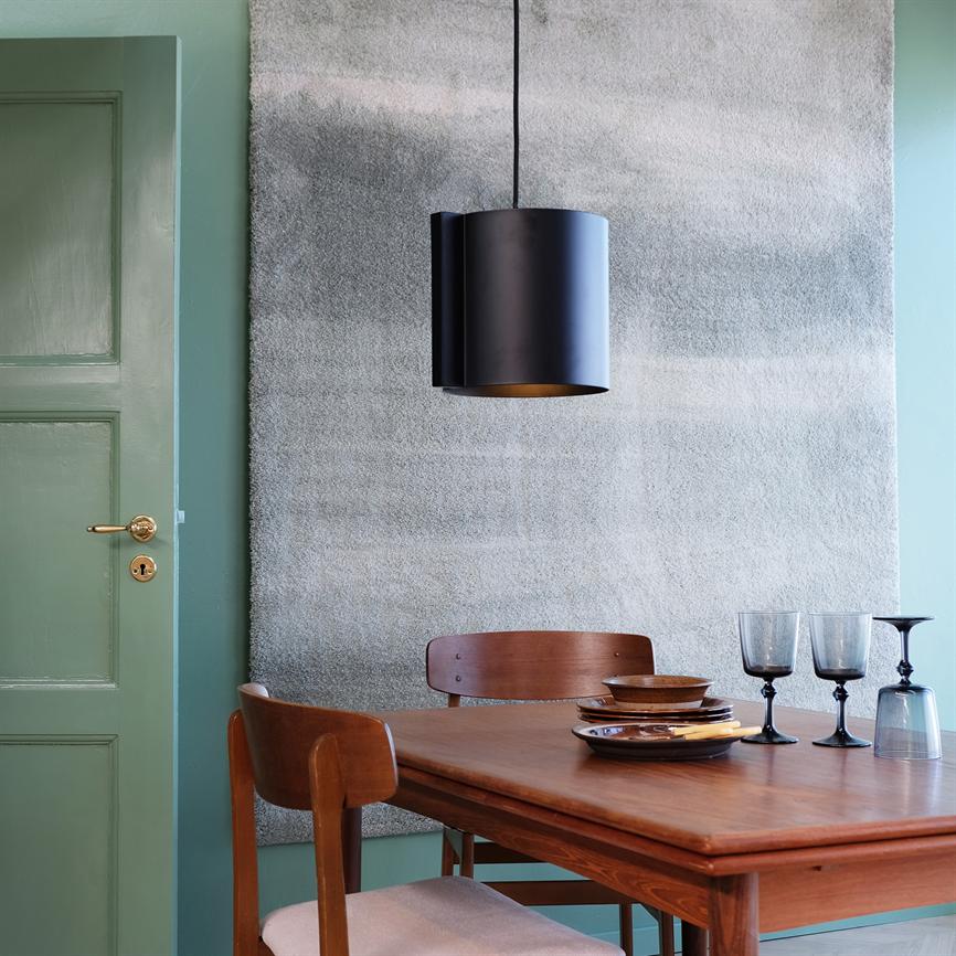 Sort cylinderformet loftlampe hænger over et teaktræsbord med stel af tallerkener og vinglas i en spisestue med mintgrøn dør, gråt vægteppe og træstole.