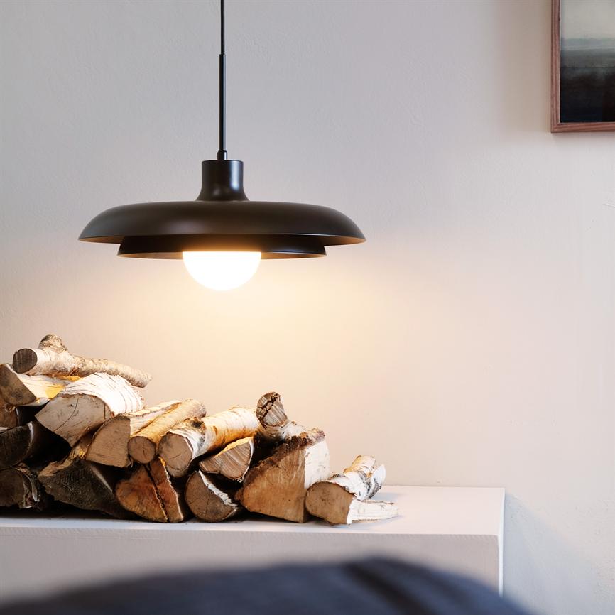 Hængelampe i sort udsender varmt, blødt lys over en bunke birkebrænde på en hvid hylde foran en lys, neutral væg.