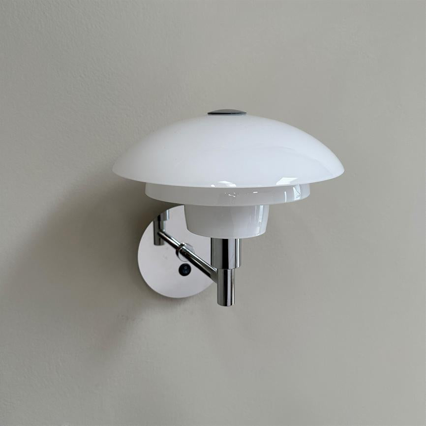 Hvid opal væglampe monteret på en blank kromarm; rund, flerlags glasskærm hæver sig over armen og sidder på en lys, ensartet beige væg.