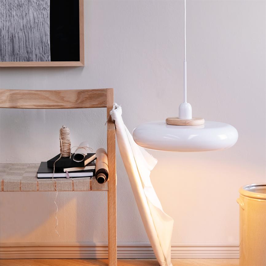 En hvid pendellampe hænger og kaster blødt lys over en træstol med bøger, snor og papirruller; et hvidt stof er draperet nedad i et lyst, minimalistisk rum.