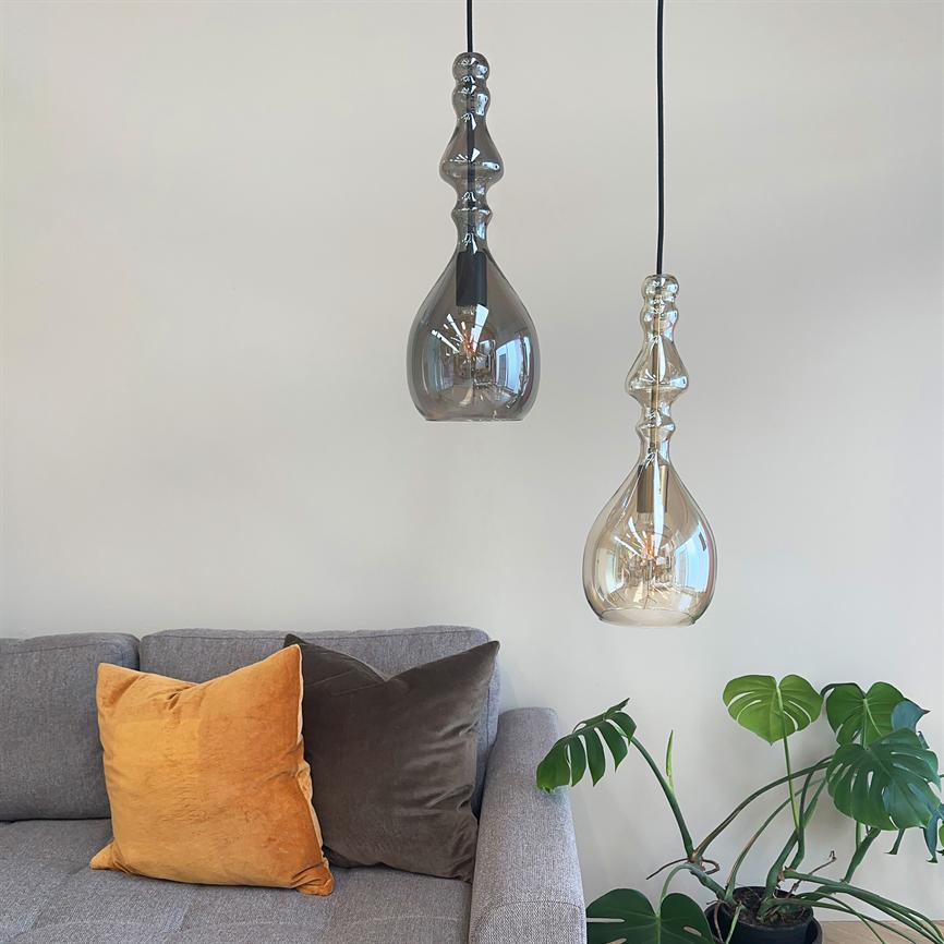 To dekorative glaspendler hænger over en grå sofa, reflekterer lys; sofaen har en gul og en brun pude, og en Monstera-plante står ved siden af mod en hvid væg.