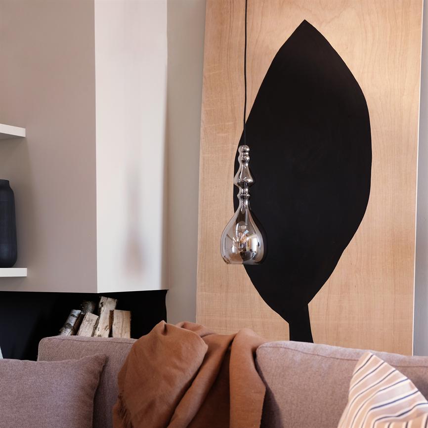 En hængende ornamenteret glaslampe reflekterer rummet foran et lyst træpanel med en stor sort bladform; i forgrunden en beige sofa med brunt tæppe og synligt brændeskur.