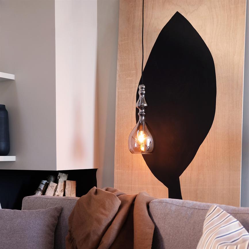 Glaspendel hænger og lyser med varmt skær foran en lys træplade med stor sort blad-silhuet; foran en grå sofa med brunt tæppe, stribet pude og stak brænde i en niche.