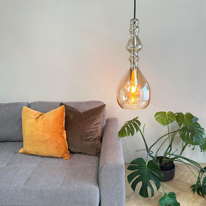 Glaspendel hænger og udsender varmt lys over en grå sofa med gul og brun pude, ved siden af en monstera i potte på lyst trægulv foran en lys væg.