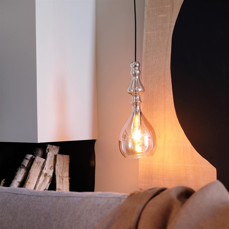 En dekorativ glaspendel hænger tændt og kaster varmt lys; ved siden af ligger stakkede birketræstammer i en niche, foran en grå sofa og abstrakt vægdekoration.