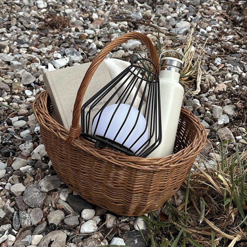 Flettet kurv rummer en kugleformet lampe i sort metalbur, en lysegrå bog og en hvid drikkeflaske, liggende på en stenfyldt strand med tørt sivgræs.