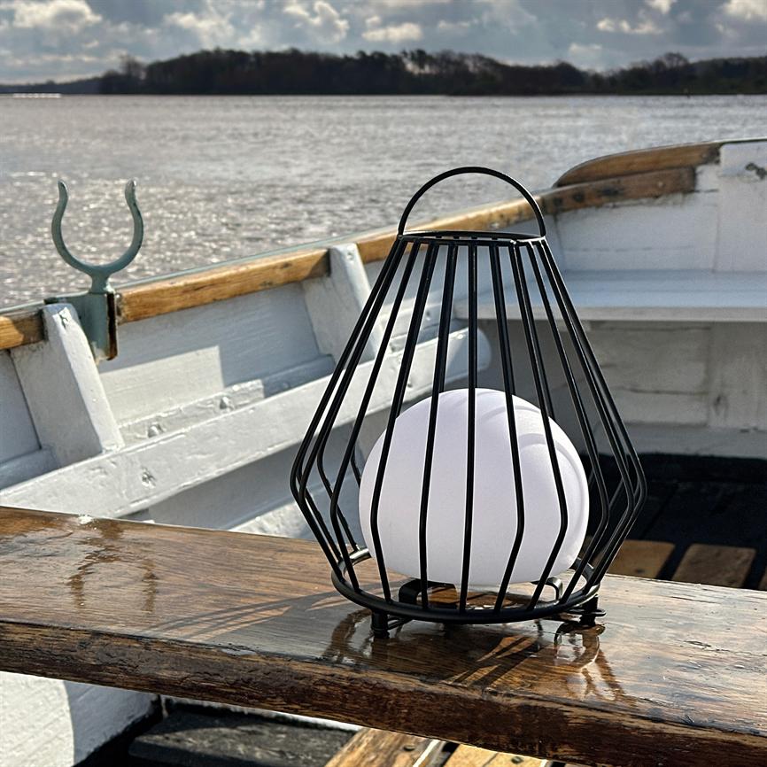 En sort metallygte indeholder en hvid kugle; står på et vådt trægelænder på en hvid robåd, foran blankt vand og en træbevokset horisont under en skyet himmel.