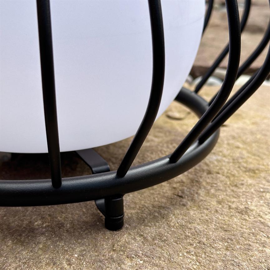 Hvid rund lampe indespærret i et sort metalbur; tynde sorte stænger omslutter globen, og lampens lille fod hviler på en ru stenflade udendørs.