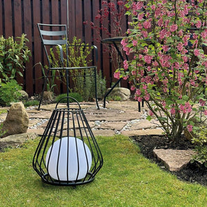 Sort metallygte med hvid kugle står på græsplænen; i baggrunden står en metalstol og et bord på en stenbelagt terrasse; til højre blomstrer en lyserød busk.