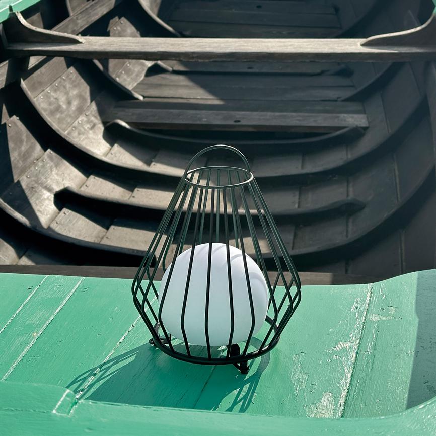 Et sort metalbur med en hvid kugle hviler på en grøn bådkant; i baggrunden ses mørke træbådsribber og skygger inde i bådens indre.
