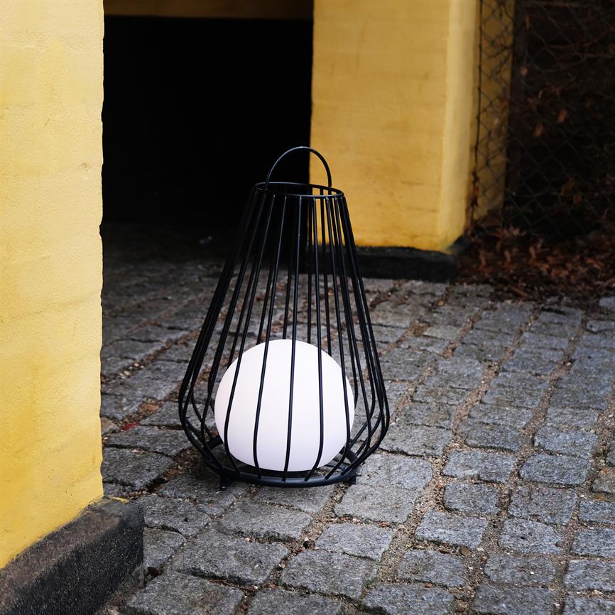 En hvid kuglelampe står i en sort gitterkurv; den lyser svagt på brosten foran en gul mur og en mørk indgang.