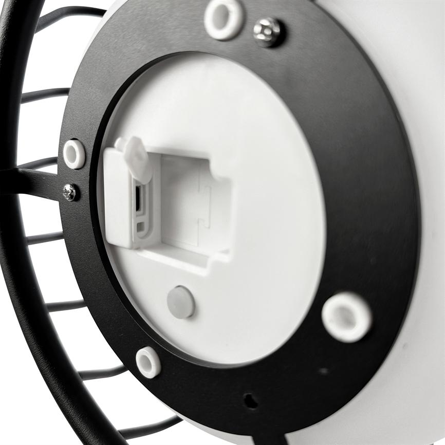 Rund sort metalramme med gitterstænger og hvid plastikindsats, der har en åben rektangulær port (mulig USB-port eller batterirum), synlige skruer og gummidutter; nærbillede mod hvid baggrund.