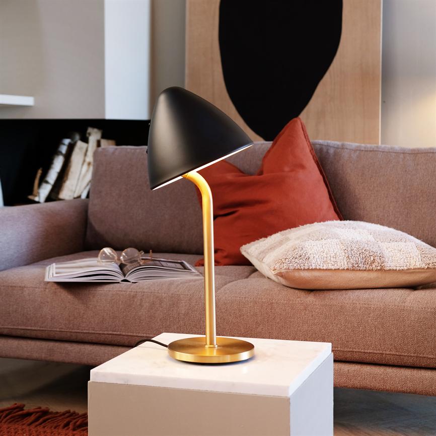 En bordlampe med sort skærm og gylden fod lyser ned på et hvidt marmorbord; foran en sofa med åben bog, briller og røde og beige puder i et hyggeligt stuehjørne.