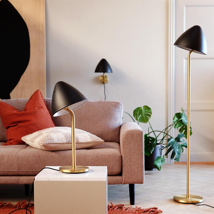 Sort og messing bordlampe lyser ned over et hvidt sidebord; foran en rosa sofa med pudebetræk og rød pude, ved siden af en gulvlampe og grøn stueplante.