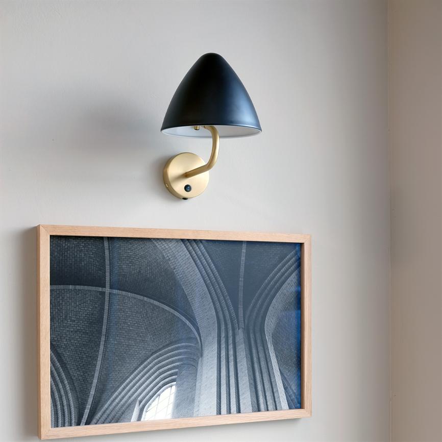 Vægslampe med sort kuppel og buet messingarm er monteret over et lyst træramt fotografi af høje, buede hvælvinger på en neutral lysegrå væg.