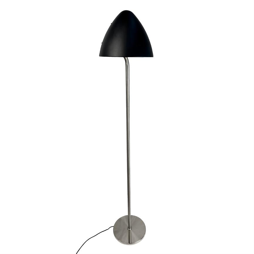Gulvlampe med sort kuppel, monteret på slank kromstang, står oprejst på en cirkulær rustfri fod med ledning, placeret foran en neutral hvid baggrund.