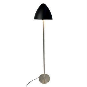 Gulvlampe står med sort kuppelformet skærm, tynd kromstang og rund metalfod; en sort ledning ligger ud fra foden mod en ensartet hvid baggrund.