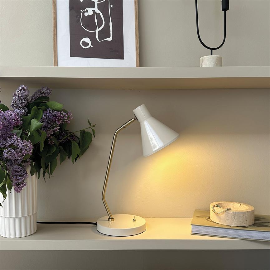 Hvid bordlampe med messingarm kaster varmt skær over en hylde med lilla syrener i hvid vase, en stentallerken på en bogbunke og en indrammet abstrakt plakat bagved.