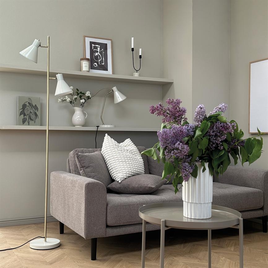 En vase med lilla syrener står på et rundt sofabord foran en grå sofa; puder, gulvlampe og væghylder med dekorative genstande giver en rolig, nordisk stuebaggrund.