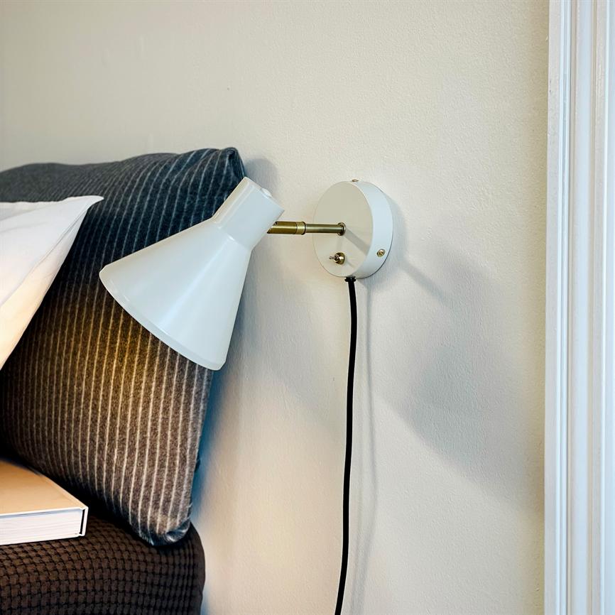 Hvid væglampe drejet nedad, fastgjort til væg med synlig sort ledning; lyser svagt mod et mørkt, stribet sengegavl ved siden af en bog på sengekanten.