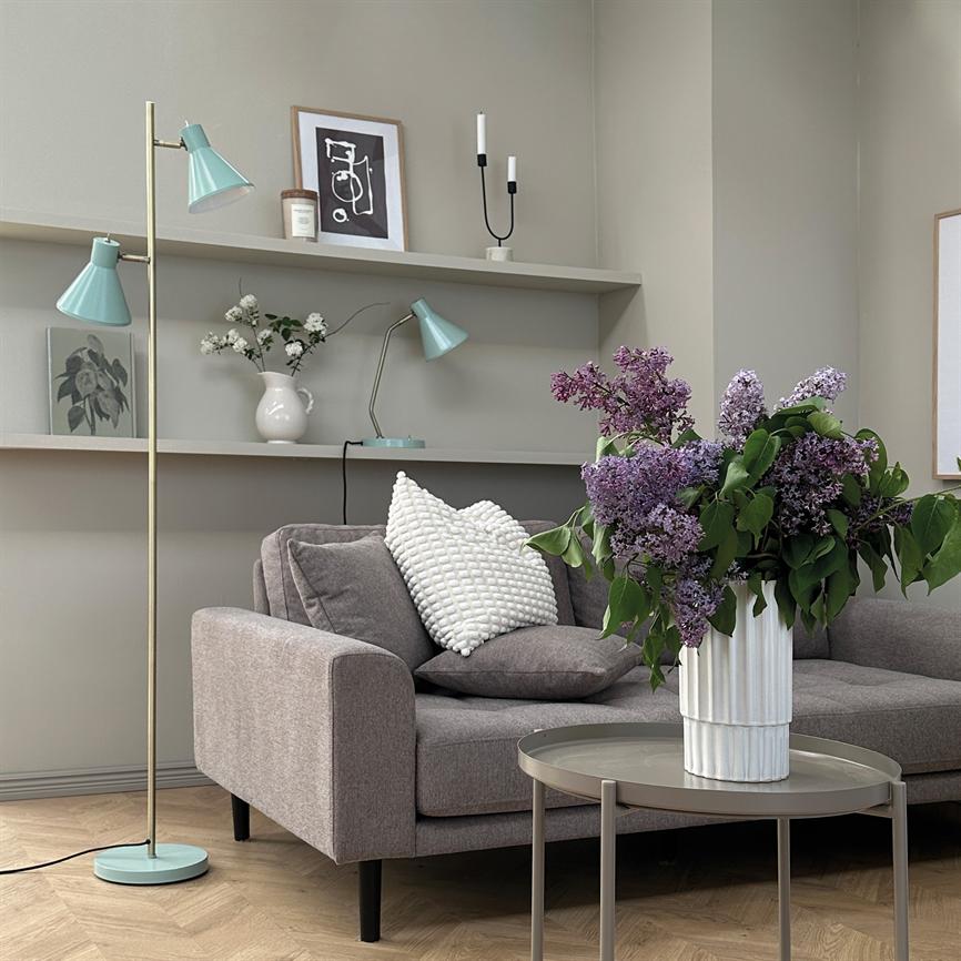 En stor buket lilla syrener i en hvid vase pryder et rundt sofabord; foran en grå sofa med pude, i et lyst, minimalistisk stuehjørne med gulvlampe og hylder.