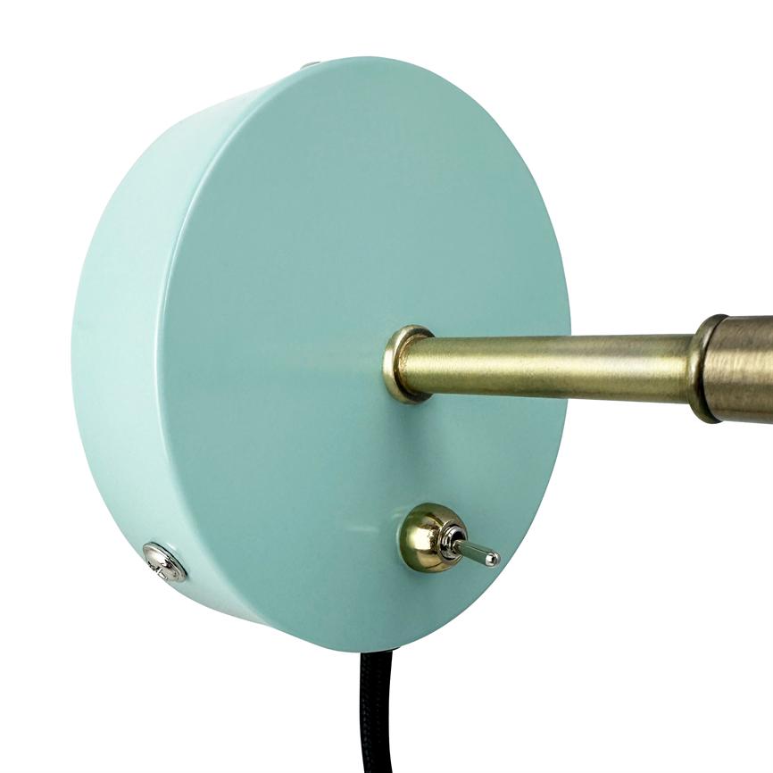 Rund mintgrøn vægplade med et messingfarvet armstykke, hvor en messingstang strækker sig ud; en lille vippekontakt stikker frem, og en sort stofbelagt ledning hænger ned mod hvid baggrund.
