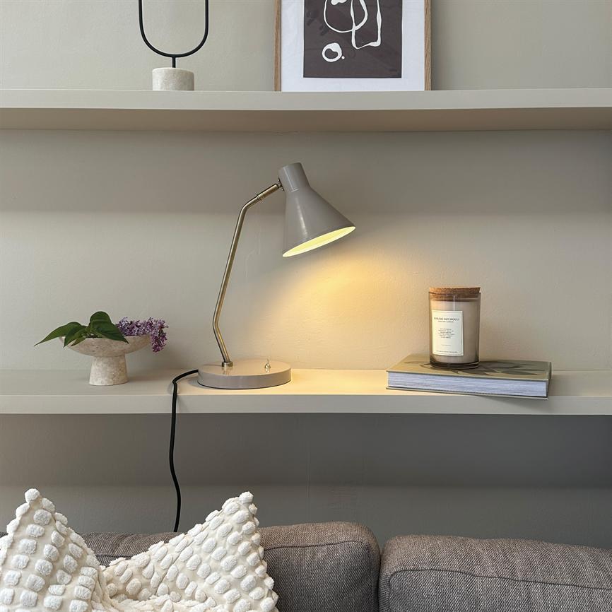 En grå bordlampe kaster varmt lys på en væghylde; den står ved en bogstak med et glasduftlys, en lille skål med blade og lilla blomster over en grå sofa med strukturerede puder. Der er tekst på duftlysets etiket, men den er for lille og uklar til at aflæse.