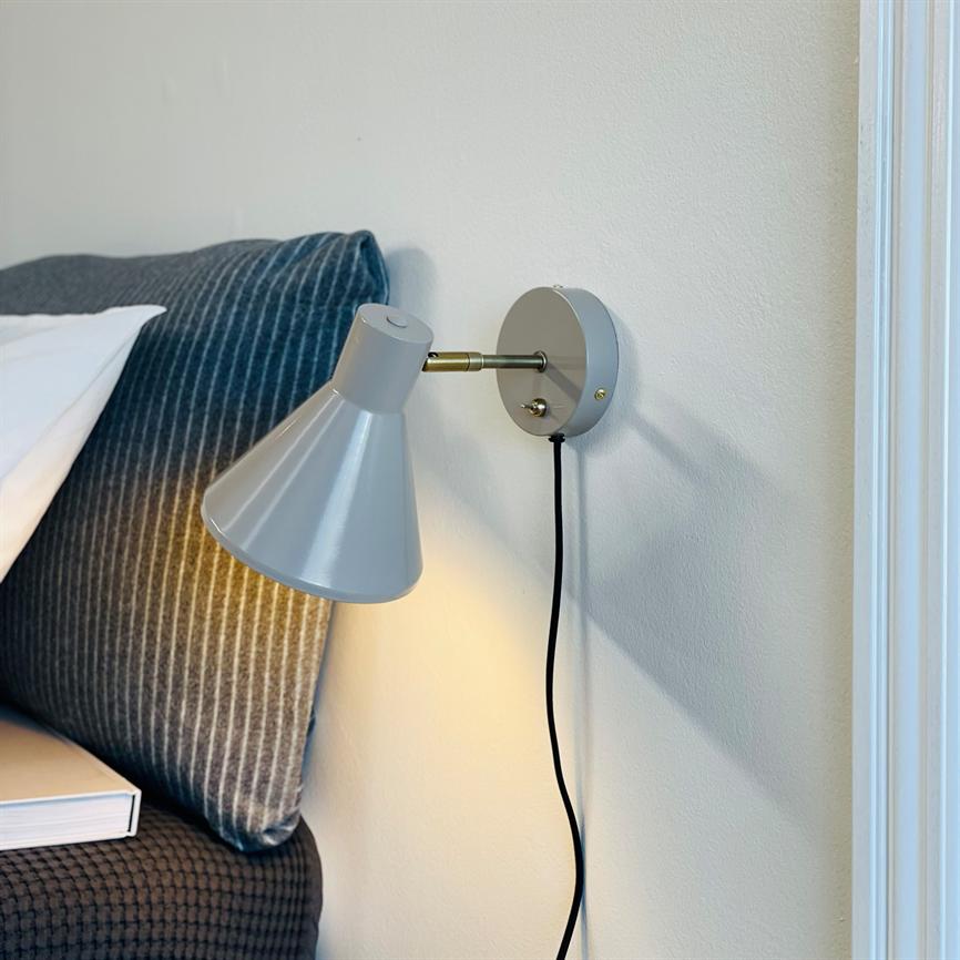 Væglampe monteret på en hvid væg lyser nedover sengegavlen; grå kegleformet skærm med sort ledning, ved siden af en mørkegrå, stribet pude og en bog på sengekanten.