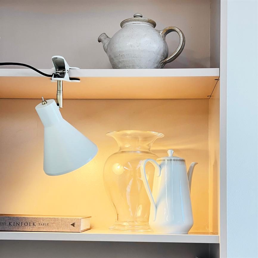 En hvid kaffekande står oplyst af en klemme-lampe; den står ved siden af en klar glasskrukke; på hylden ovenover en grå keramiktekande og nederst en bog. Tekst på bogryg: "THE KINFOLK TABLE".