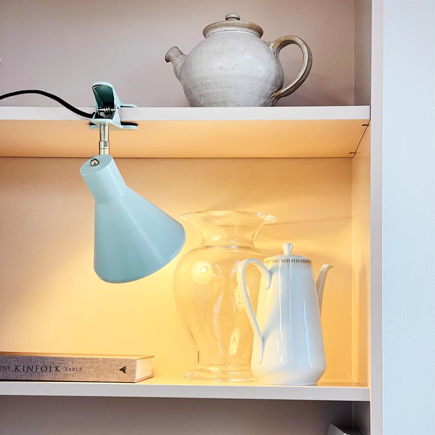 En mintgrøn klemme-lampe lyser på en klar glaskrukke og en hvid kande på en oplyst hylde, under en keramisk tekande; bogryg med teksten THE KINFOLK TABLE.