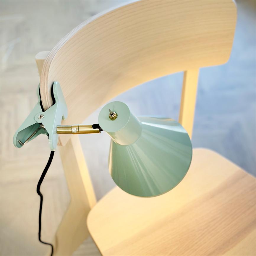 Klemme-lampe i mintgrøn klamrer sig fast på ryggen af en lys træstol, lyser ned mod sædet; sort ledning hænger, lyst trægulv ses i baggrunden.