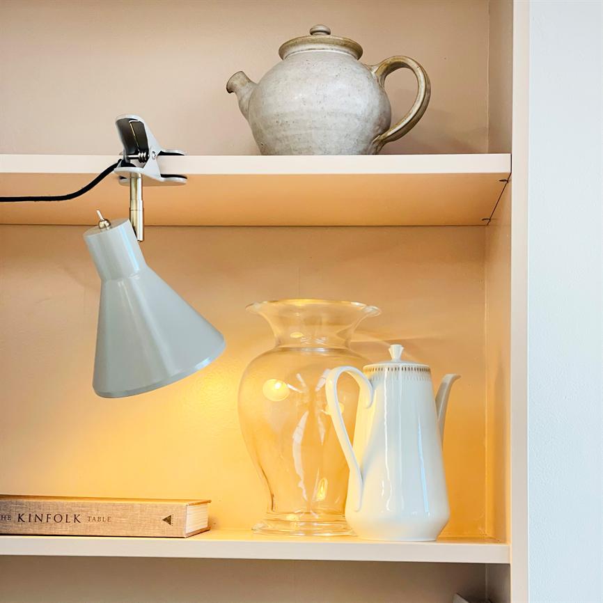 Hvid porcelænsk kaffekande står ved siden af en klar glasvase, oplyst af en grå klemme-lampe, på en lysebeige indbygget hylde med en lertekande øverst. 
 Tekst: THE KINFOLK TABLE